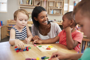 Three young children and one caretaker in a daycare room playing with a puzzle game.